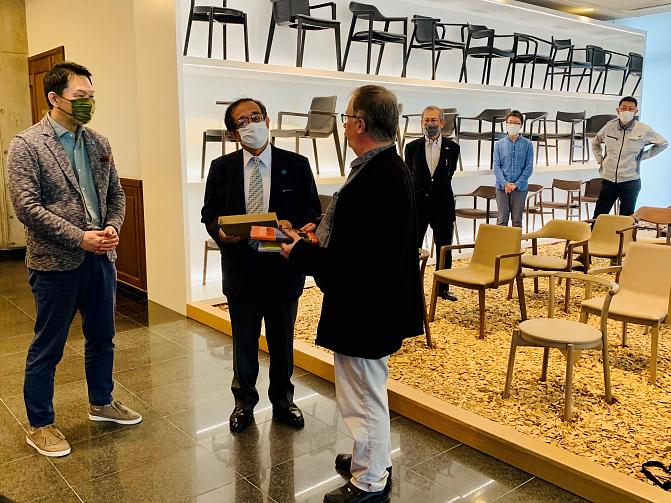 Three people are having a conversation at a japanese furniture factory, there are rows of chairs in the background.