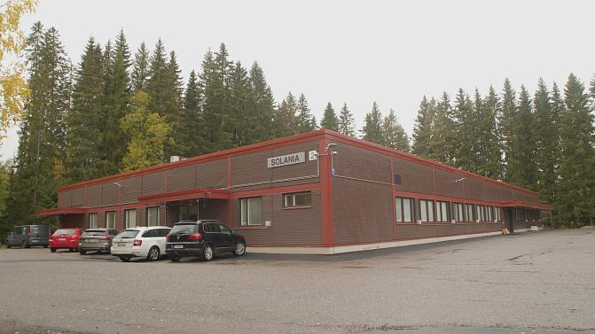 Luke Jokioisten ruskea Solania-rakennus. Vasemmalla seinustalla parkissa autoja, taustalla metsää.