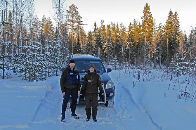 A police officer and a game warden are standing in front of a car in the forest.