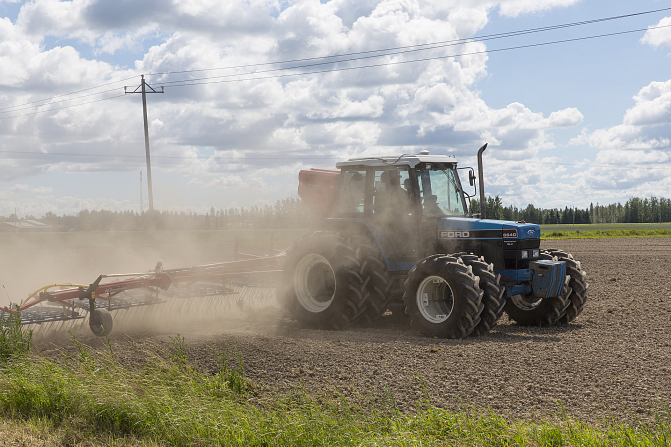 Tractor is driving on a field. It is early summer and there are white clouds on the blue sky.