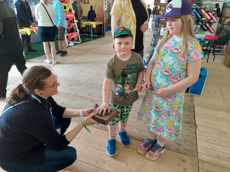 Lapsi tunnustelee suden tassunjäljestä tehtyä pronssivalosta, jota kyykistynyt aikuinen pitelee. Toinen lapsi katselee vieressä.