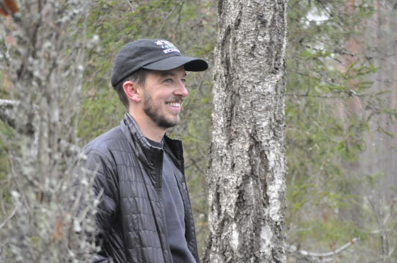 A smiling man wearing a cap standing among trees in the forest.