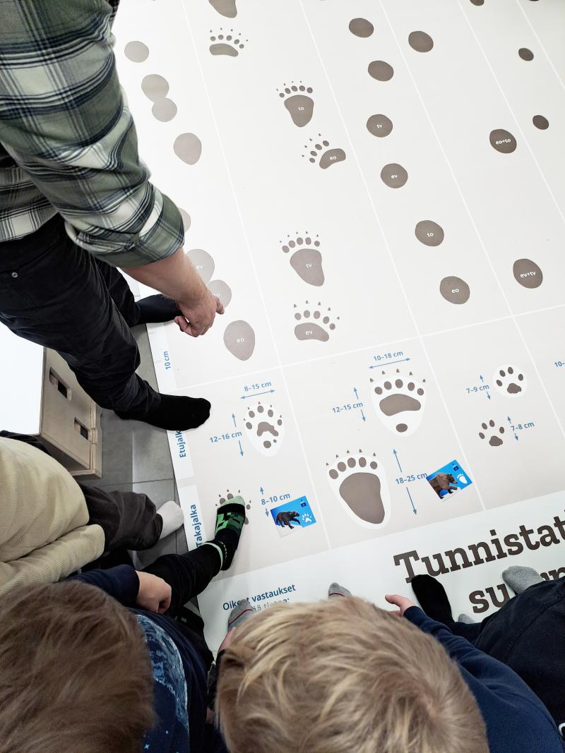 Koululaisia ja kouluvierailija seisovat jälkimatolla, jossa näkyy oikean kokoisia petojen jälkiä. Matolle on laitettu pedoista kertovia tarroja.