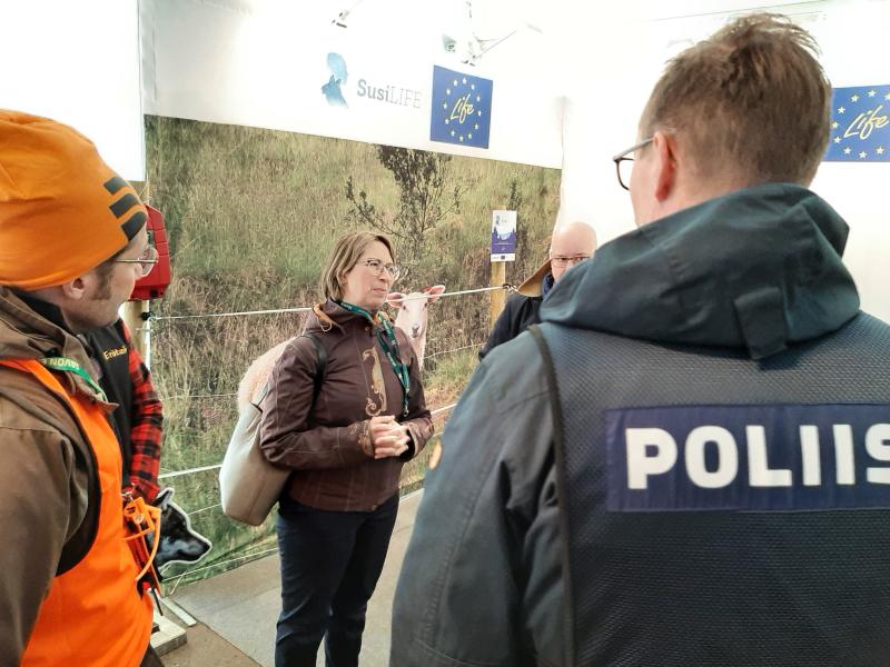 I bilden syns en mässmonter där ett rovdjursstängsel har rest. I mässmontern diskuterar en grupp människor. En av personerna är minister Sari Essayah. I förgrunden av bilden står en äldre konstapel i polismundering och en jägare i orange varselfärg, både med ryggen mot fotografen.