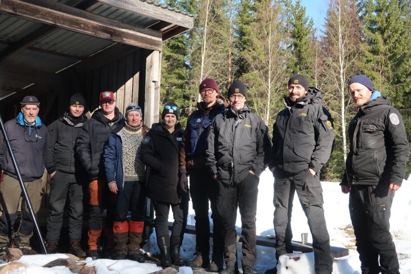 Ulkona kevättalven hangessa laavun edessä seisoo yhdeksän ihmistä hymyilemässä kameralle.