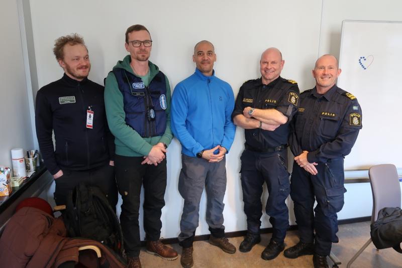 Viisi miestä seisoo kokoushuoneessa ja hymyilee kameralle. Kaksi miehistä ovat pukeutuneet Ruotsin poliisin univormuun, yhdellä on Suomen poliisin liivi ja yhdellä erätarkastajan työvaatteet.