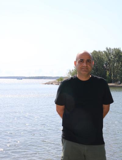 Khazaei in a medium shot standing outside by the shore in the sun. He looks straight to the camera and smiles. 
