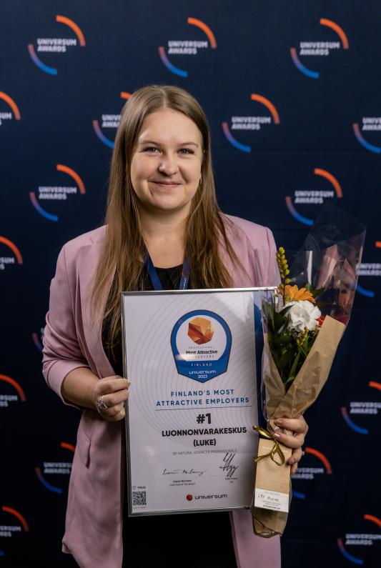 Luken HR-asiantuntija Linda Yli-Keturi katsoo kameraan kädessään todistus ykkössijasta ja kukkakimppu. 
